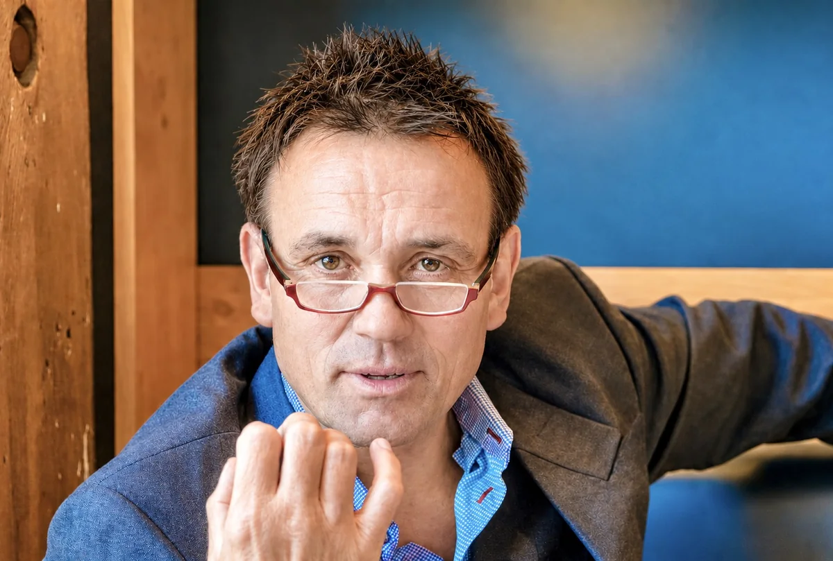 Lukas Scheja mit roter Brille im Gespräch, eine Hand erhoben, vor Holzwand und blauer Wand.