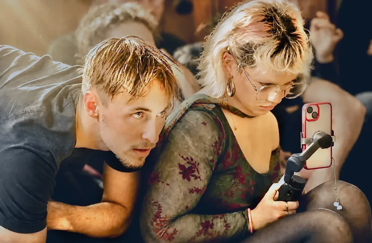 Zwei junge Schauspielende dicht nebeneinander, eine Person hält ein Smartphone an einem Stabilisator.