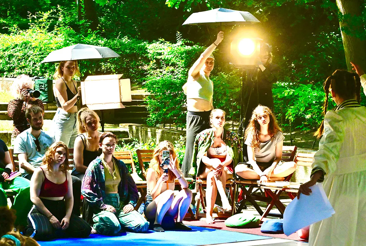 Sommerakademie im Freien, junge Menschen sitzen im grünen Park vor Filmlicht und Softboxen und verfolgen eine Szene im Sonnenlicht