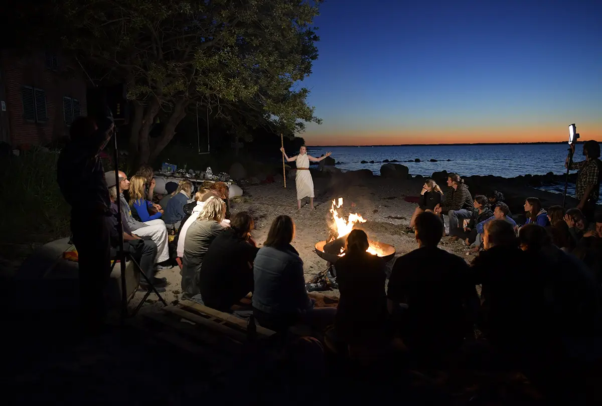Ensemble der Sommerakademie sitzt abends am Lagerfeuer am Ufer, im Hintergrund der Sonnenuntergang über dem Wasser