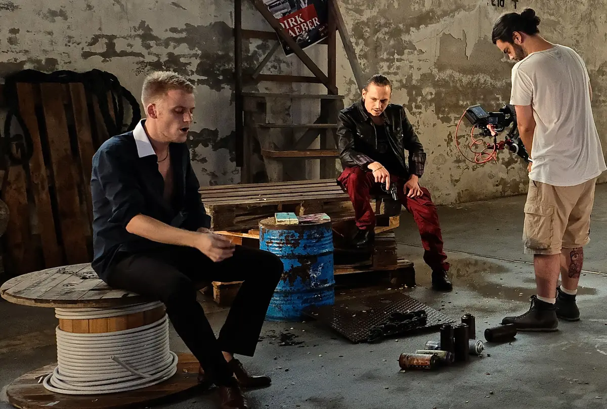 Zwei Darsteller in einer Szene in einer verlassenen Halle, daneben ein Kameramann mit der Filmkamera im Schultergriff.