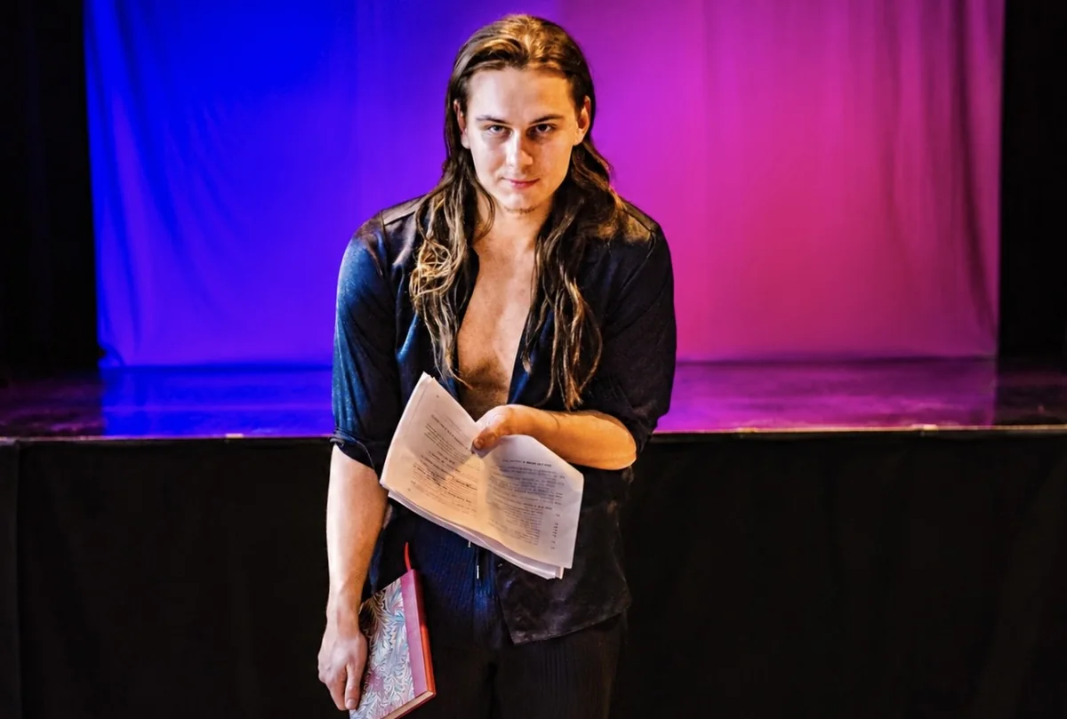 Schauspieler steht mit aufgeschlagenem Textbuch auf der Bühne, hinter ihm violettes und blaues Licht.