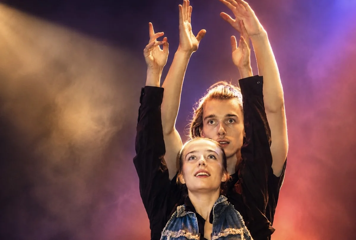 Zwei Schauspielende strecken im bunten Bühnenlicht die Arme nach oben und blicken nach oben.