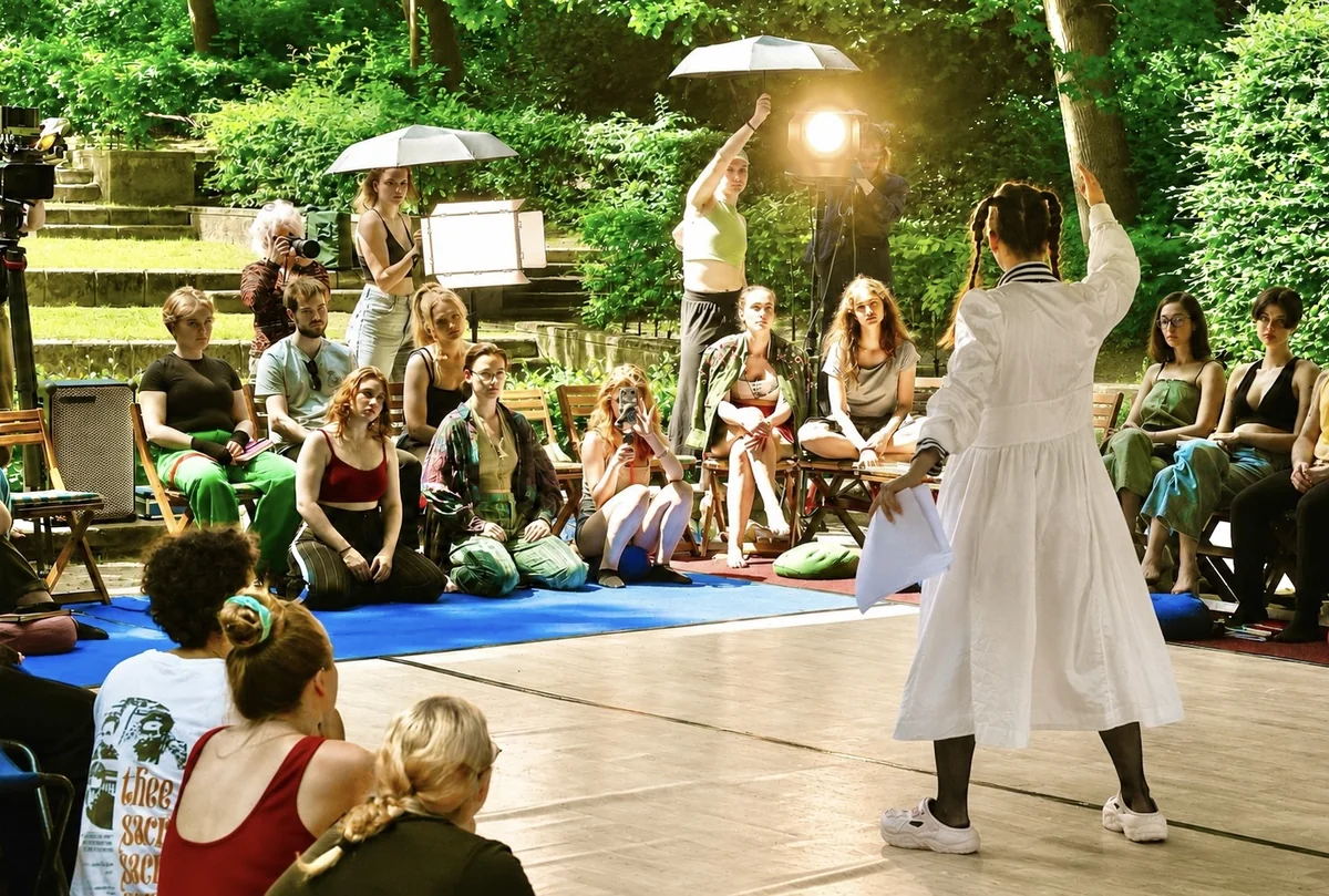 Sommerakademie im Park, eine Darstellerin steht mit erhobenem Arm vor der sitzenden Gruppe, daneben Scheinwerfer und Kamera.