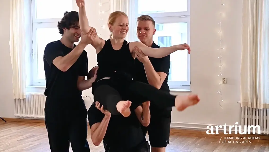 Vier Teilnehmende im Schauspielworkshop, drei tragen lachend eine Person waagerecht durch den hellen Proberaum mit Holzboden und Fenstern