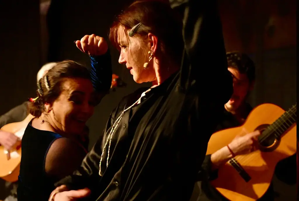 Flamenco-Szene: eine Tänzerin mit erhobenem Arm, daneben eine weitere Frau, im Hintergrund Gitarristen.