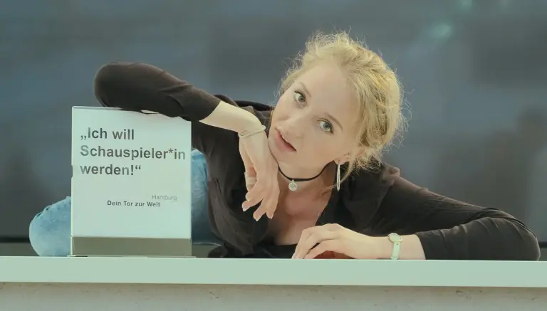 Junge blonde Frau lehnt neben einem Schild mit der Aufschrift „Ich will Schauspieler*in werden!".