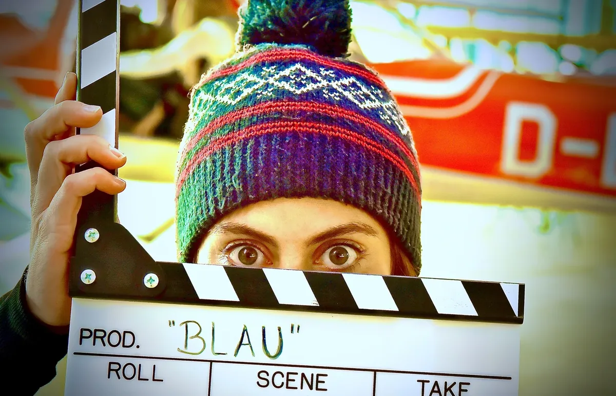 Junge Schauspielerin mit bunter Strickmütze schaut mit großen Augen über eine Filmklappe mit der Aufschrift „BLAU“