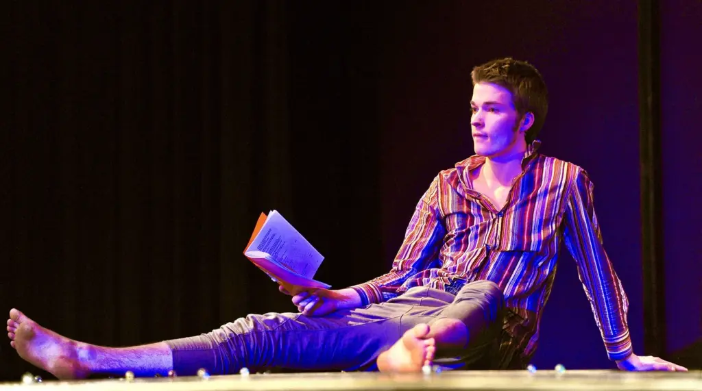 Ein junger Schauspieler sitzt auf einer Bühne und hält ein Buch in der Hand.