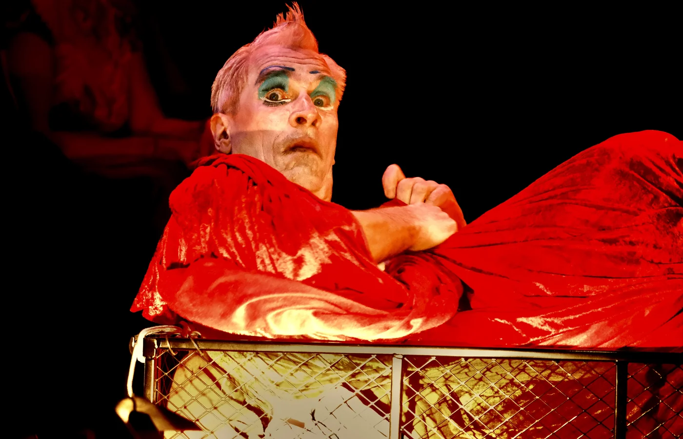Expressiver Butoh-Auftritt, eine Person mit rotem Irokesen, gruener Augenschminke und weiss geschminktem Gesicht, in rotes Samttuch gehuellt vor dunkler Buehne