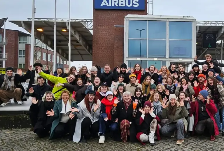 Große Gruppe Artrium-Studierender winkt gut gelaunt vor dem Eingang eines Airbus-Werksgeländes.