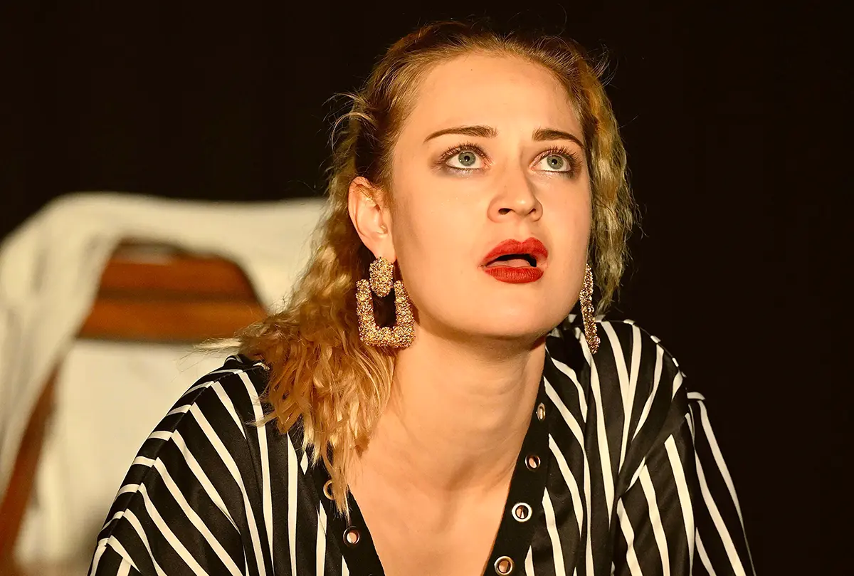 Schauspielerin mit blondem Haar und großen goldenen Ohrringen in gestreiftem Oberteil, mit aufgerissenen Augen in einer intensiven Bühnenszene