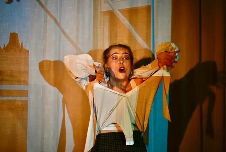 Junge Schauspielerin auf der Bühne in einer expressiven Pose mit weit geöffnetem Mund und ausgestreckten Armen, dramatisches Licht wirft Schatten an die Wand.