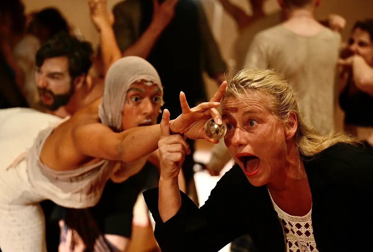Intensive Butoh-Szene, eine Frau mit weit geoeffnetem Mund haelt eine Glaskugel vors Auge, dahinter Teilnehmende in Bewegung
