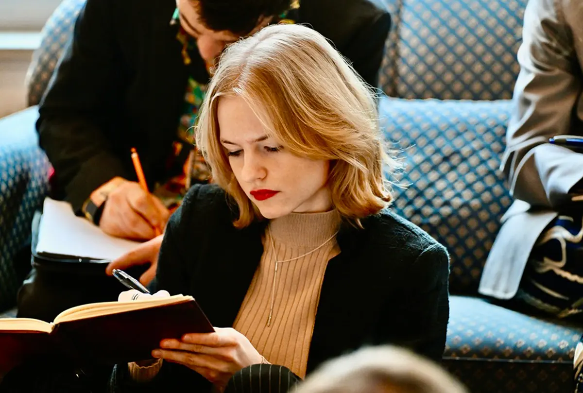 Blonde Studentin liest konzentriert in einem Notizbuch, dahinter eine schreibende Person auf einem blauen Sofa