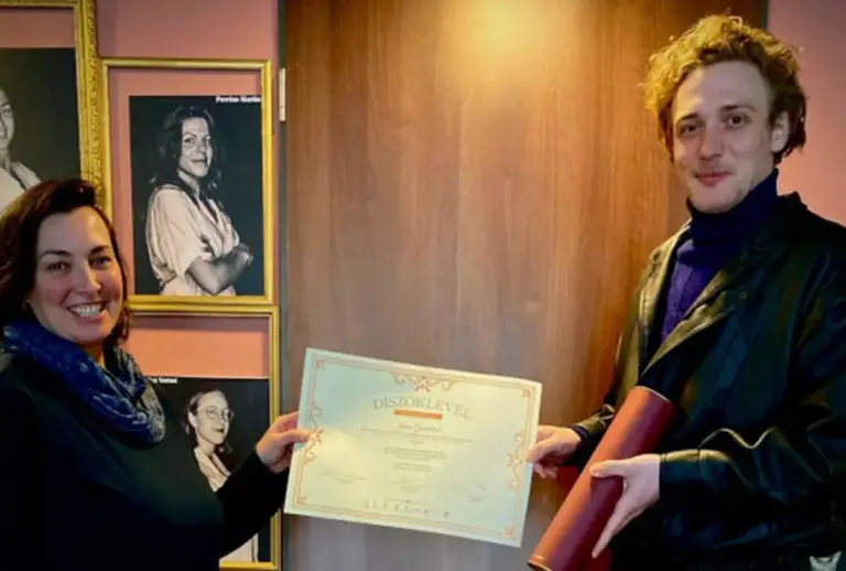 Eine Frau und der junge Schauspieler Jonas Gundlach halten gemeinsam eine Urkunde vor einer Wand mit gerahmten Porträts.