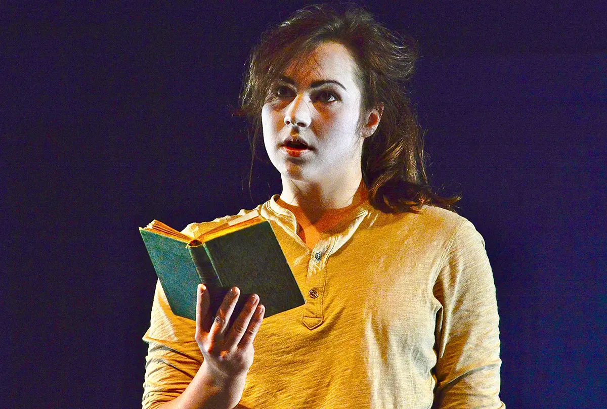 Schauspielerin mit weiß geschminktem Gesicht hält ein aufgeschlagenes Buch im warmen Bühnenlicht.