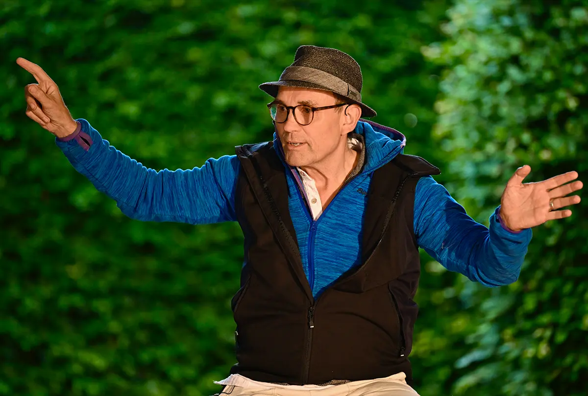 Lukas Scheja mit Hut und Brille spielt gestikulierend eine Szene vor einer grünen Hecke
