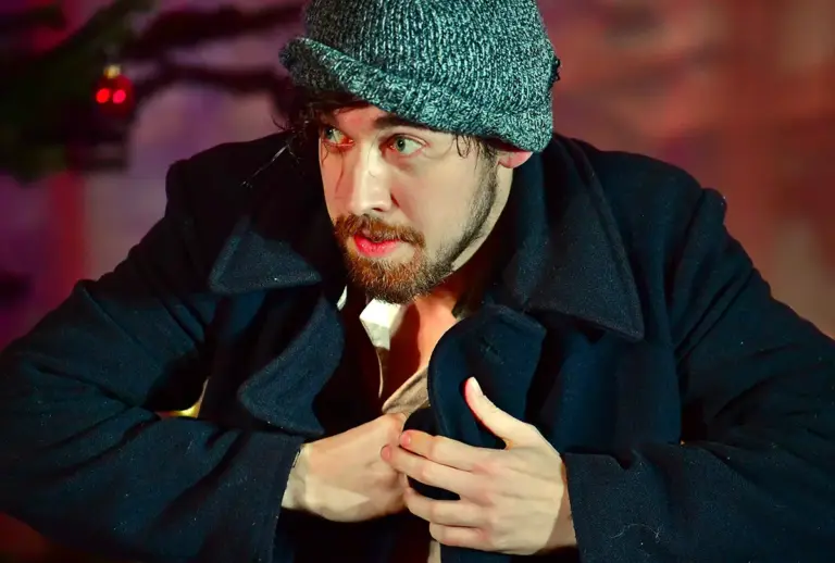 Junger Schauspieler in einer Bühnenszene, mit Wollmütze und dunklem Mantel, greift angespannt an seinen Kragen.