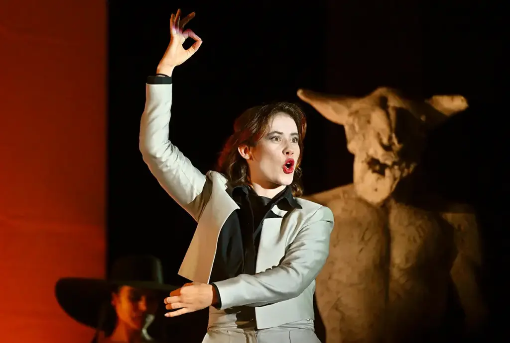 Schauspielerin in hellgrauem Anzug mit schwarzem Hemd, den Arm erhoben und im Ausruf, auf der Bühne neben einer Stierfigur.
