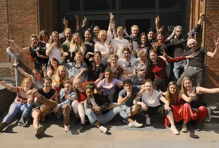 Große Gruppe fröhlicher Artrium-Studierender posiert vor der Backsteinfassade der Schule.