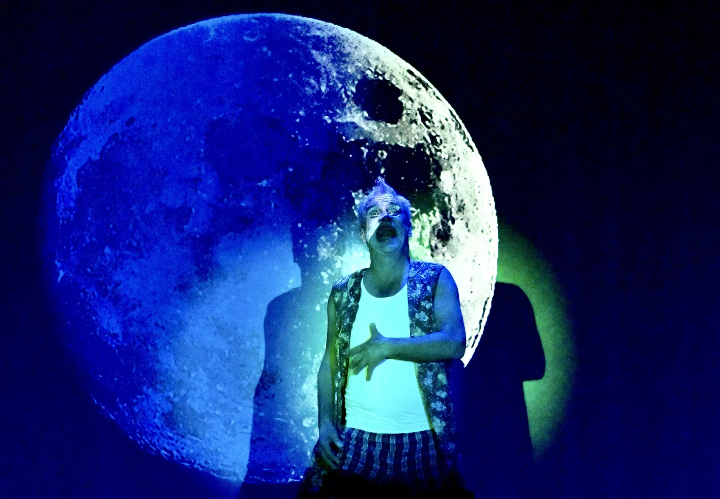 Ein Darsteller steht im blauen Bühnenlicht vor einem riesigen Mond und blickt mit geöffnetem Mund nach oben.
