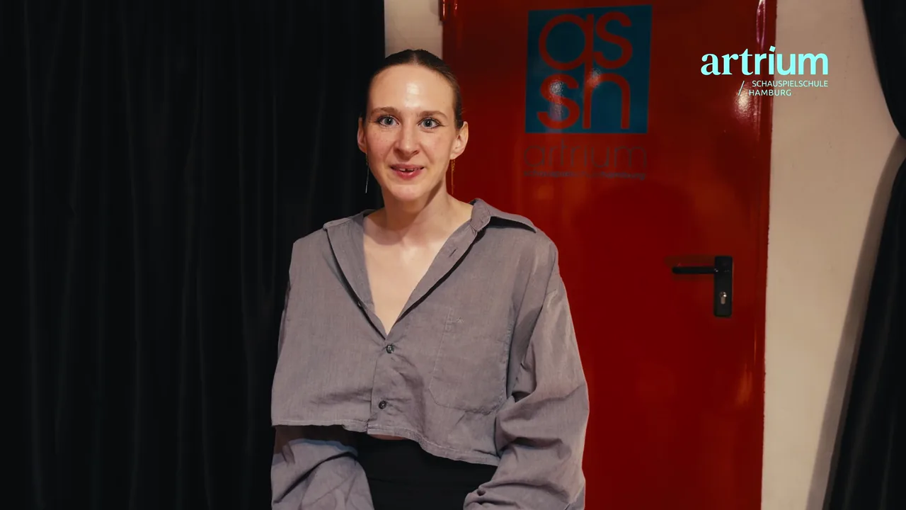 Julia lächelt vor der roten Tür mit dem Artrium-Logo und erzählt von ihrer berufsbegleitenden Schauspielausbildung.