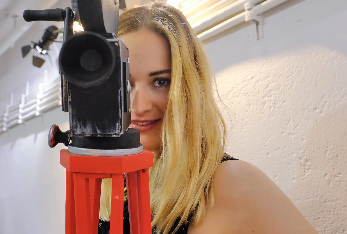Junge Frau steht hinter einer alten Filmkamera auf rotem Holzstativ und blickt lächelnd daran vorbei.