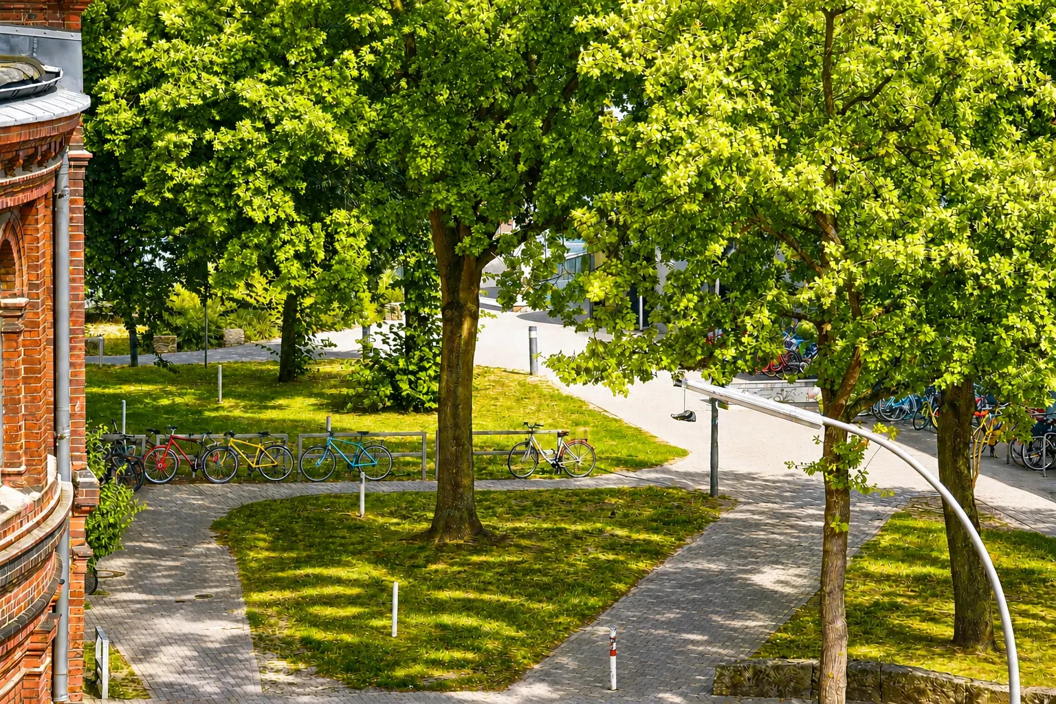 Grünfläche neben dem Artrium mit Wegen, Fahrradbügeln und alten Bäumen, links die Rundung des Backsteinbaus