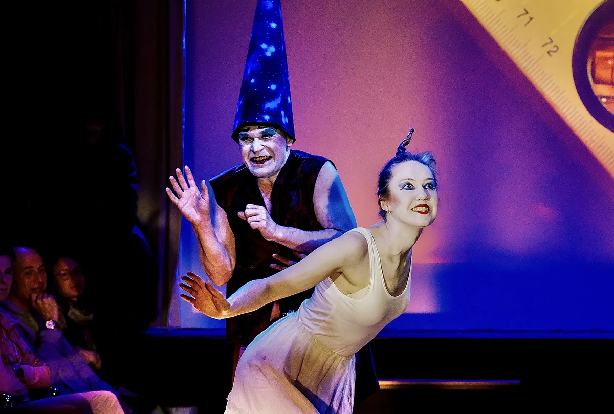 Zwei Schauspielende in einer Bühnenszene, er mit hohem Sternenhut und weiß geschminktem Gesicht, sie im hellen Kleid nach vorn gebeugt, im violetten und blauen Bühnenlicht.
