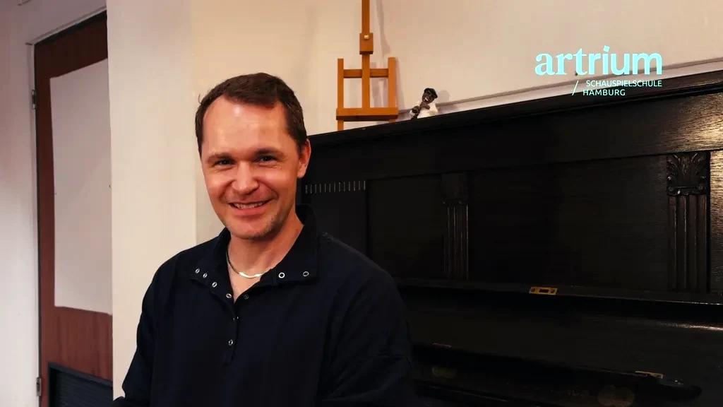 Johannes sitzt lächelnd neben einem Klavier und erzählt von der berufsbegleitenden Ausbildung am Artrium