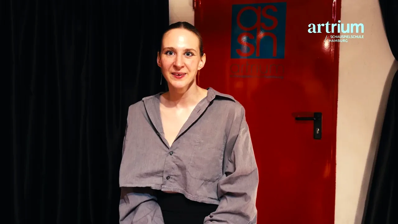 Julia lächelt vor der roten Tür mit dem Artrium-Logo und erzählt von ihrer berufsbegleitenden Schauspielausbildung.