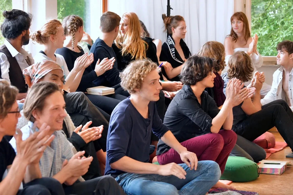 Große Gruppe Studierender sitzt im hellen Unterrichtsraum auf dem Boden und applaudiert