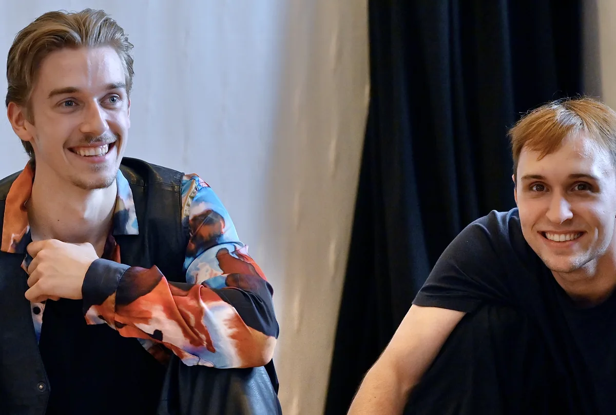 Zwei junge Schauspielstudierende sitzen im Probenraum und lachen in die Kamera