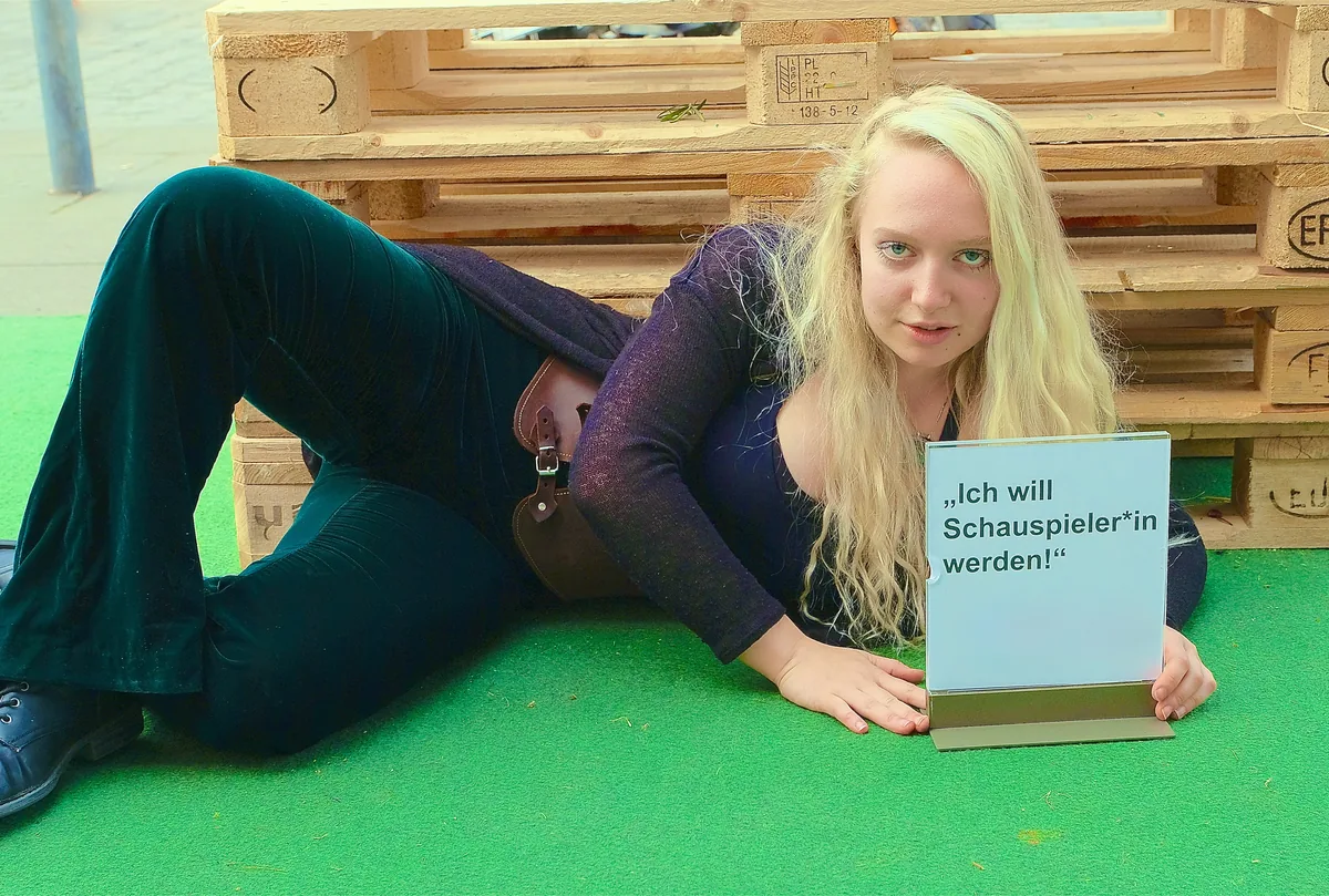 Junge Frau liegt auf grünem Boden und hält ein Schild mit der Aufschrift „Ich will Schauspieler*in werden!“
