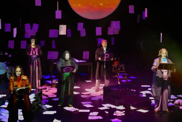 Das Ensemble mit weißen Masken und Tafeln auf der Bühne, im Hintergrund eine blutrote Sonne