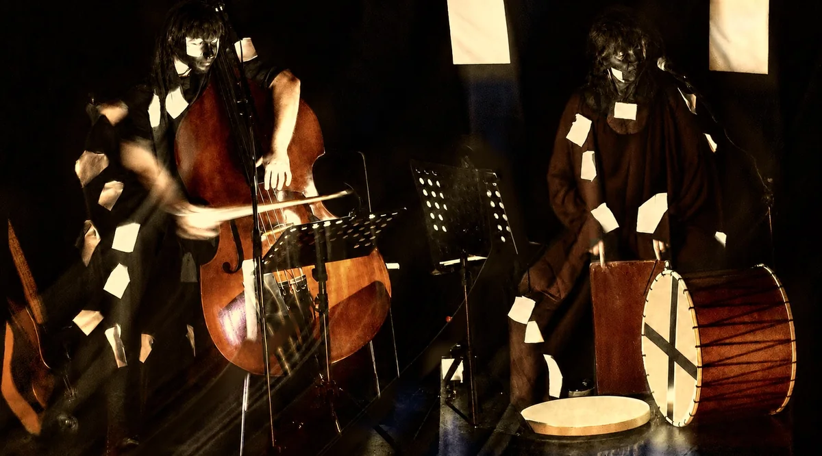 Die beiden Musiker mit Kontrabass und Trommel zwischen fallenden Papierseiten