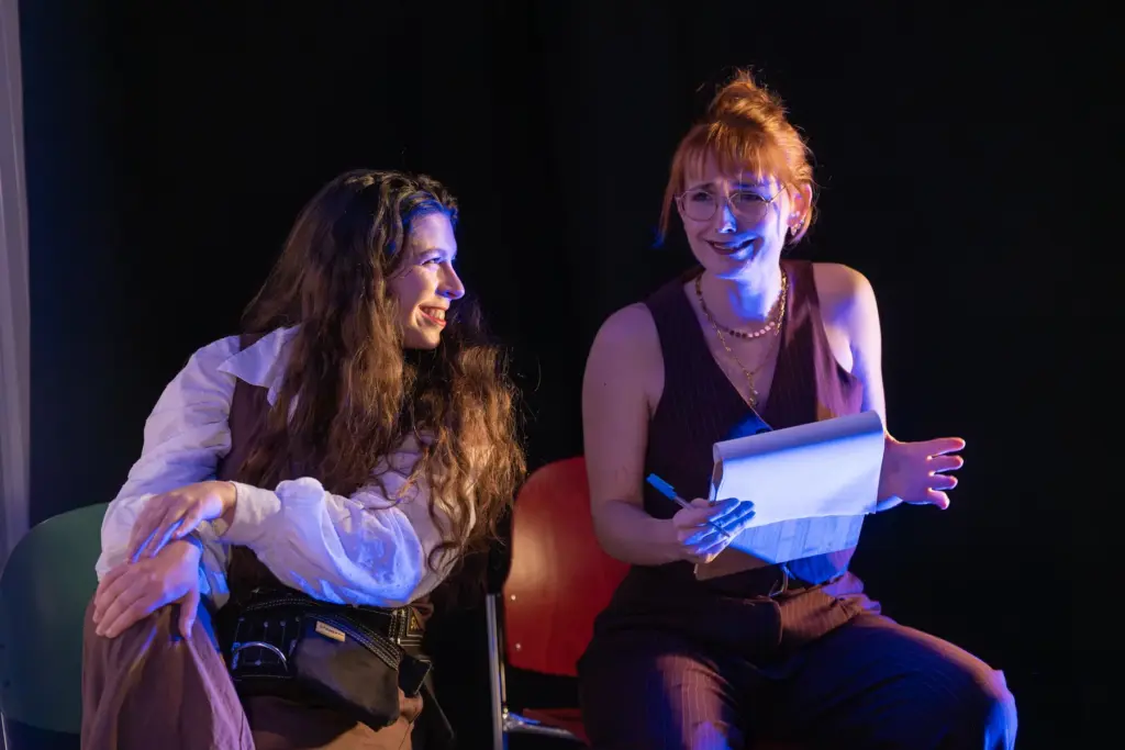 Zwei Schauspielerinnen des MUSE Kollektivs sitzen lachend auf der Bühne, eine hält ein Textbuch in der Hand