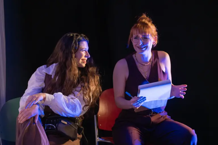 Zwei Schauspielerinnen des MUSE Kollektivs sitzen lachend auf der Bühne, eine hält ein Textbuch in der Hand