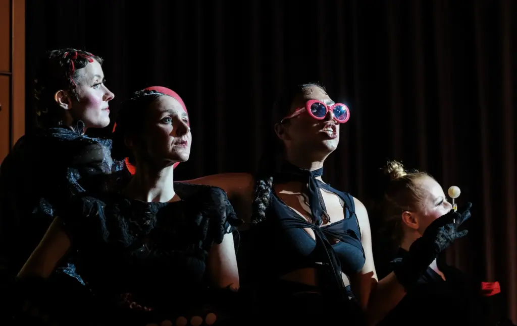 Vier Schauspielerinnen in schwarzen Kostümen blicken nach oben, eine trägt eine rosa Brille