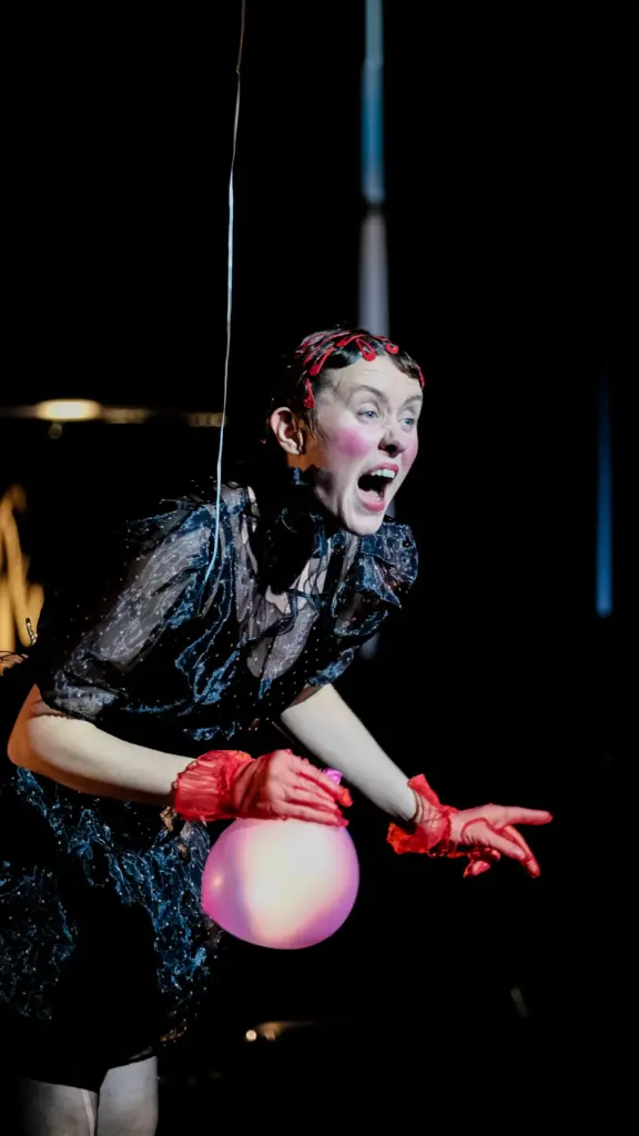 Schauspielerin in schwarzem Spitzenkostüm und roten Handschuhen mit einem rosa Luftballon auf der Bühne