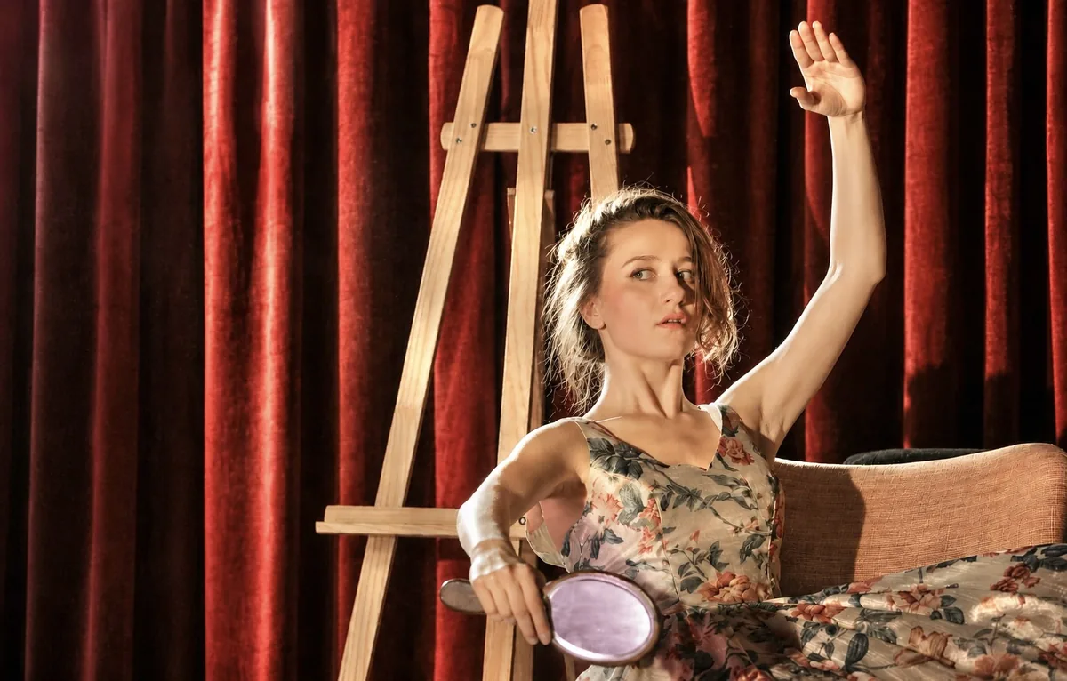 Eine Schauspielerin im geblümten Kleid sitzt vor einem roten Vorhang in einem Sessel, hebt einen Arm und hält einen Handspiegel.