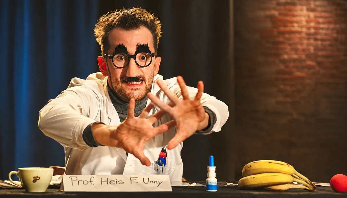 Ein Schauspieler mit Scherzbrille und Schnurrbart spielt einen Professor im Kittel und streckt beide Hände nach vorn, vor ihm liegen Bananen und eine rote Clownsnase.