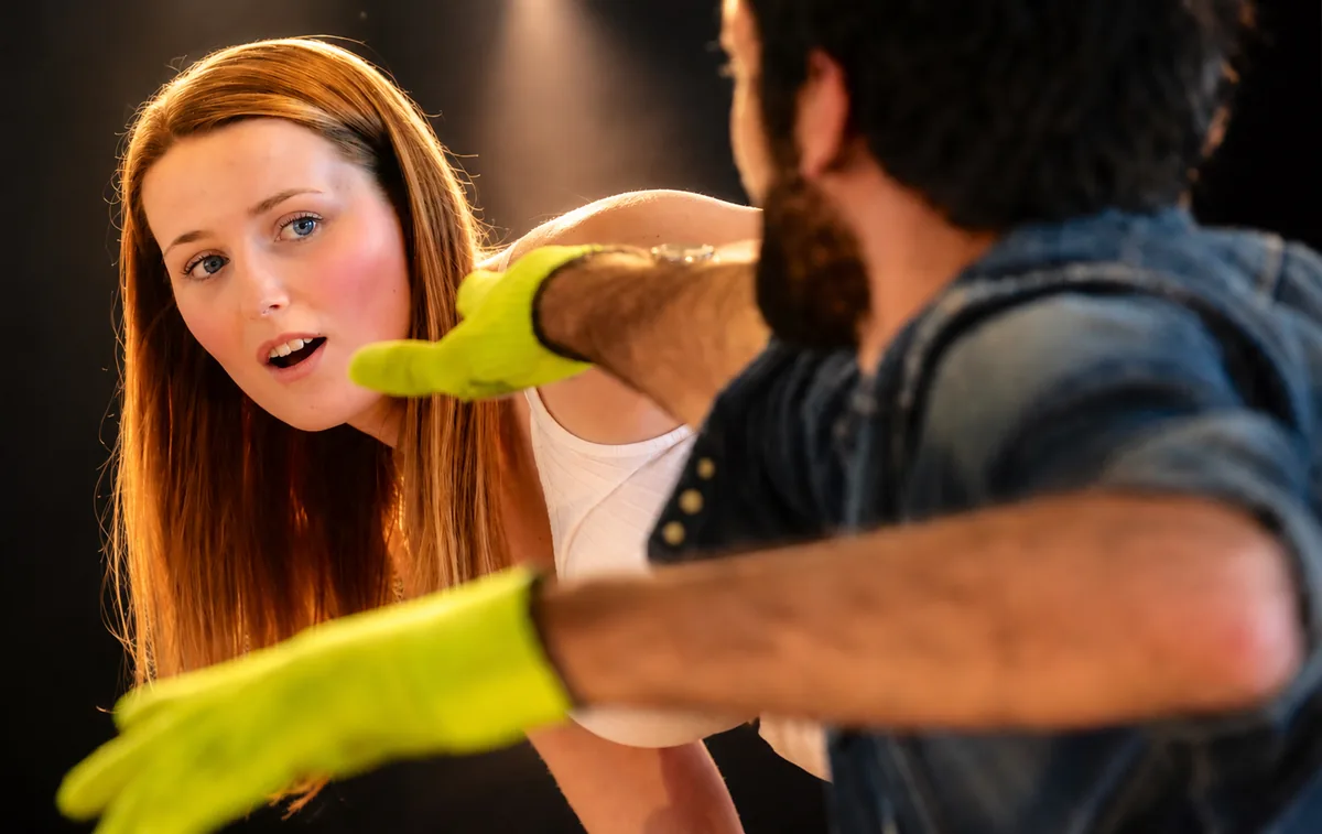 Eine junge Frau blickt in einer Szene einem Mann mit gelben Gummihandschuhen entgegen, der ihr die Arme entgegenstreckt