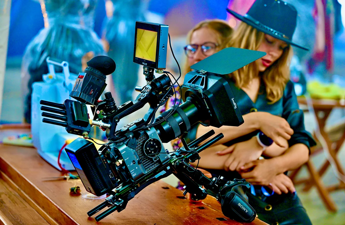 Eine Filmkamera mit Monitor steht im Vordergrund auf einem Holztisch, dahinter stehen zwei junge Frauen an einem Filmset.