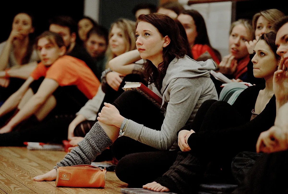 Schauspielschülerinnen und Schauspielschüler sitzen im Unterricht auf dem Probenboden und hören konzentriert zu