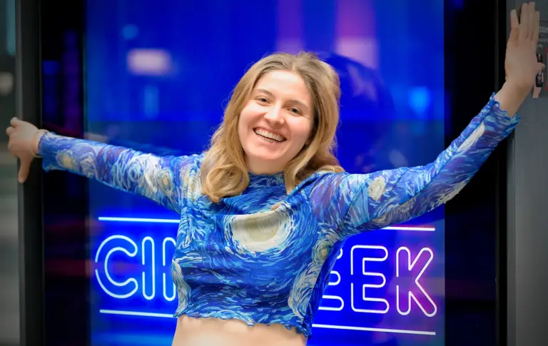 Maria Hein, Studierende am Artrium, lacht mit ausgebreiteten Armen vor einer blau leuchtenden Neonschrift